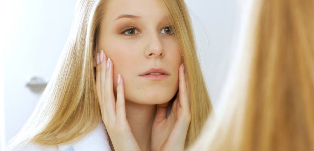 Young blonde woman checking skin in mirror